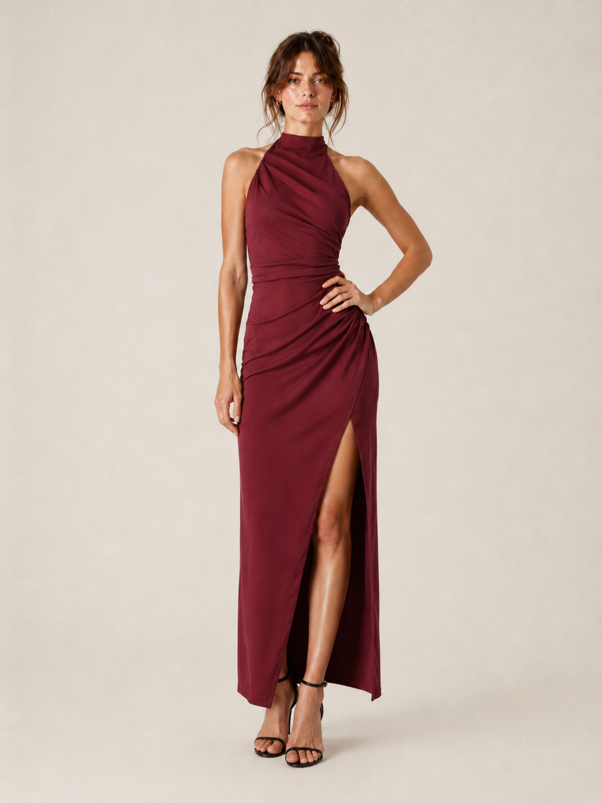 A woman viewed from a front three-quarter angle wearing the Elévaison Maren burgundy ruched halter-neck maxi dress with a side slit, against a light beige studio background.