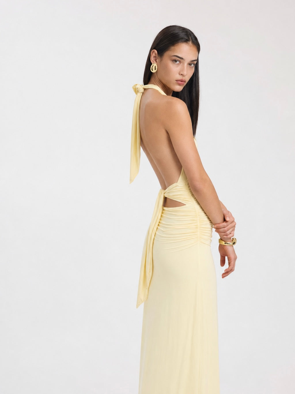 A woman viewed from a back three-quarter angle wearing the Elévaison Vivienne light yellow deep V-neck halter maxi dress with a ruched waist, against a light gray studio background.