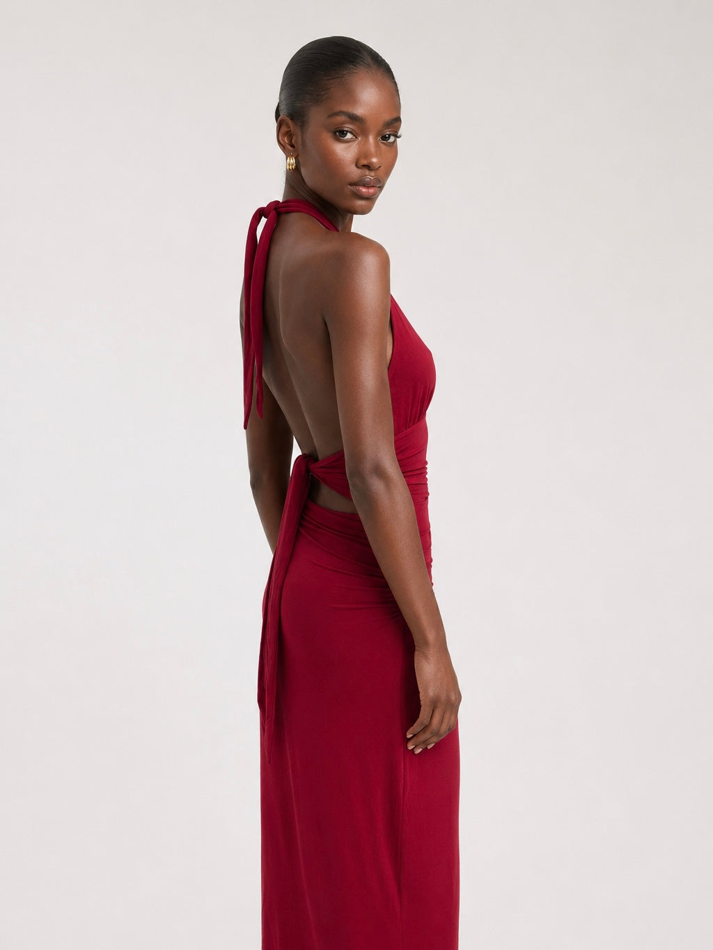 A woman viewed from a back three-quarter angle wearing the Elévaison Vivienne burgundy deep V-neck halter maxi dress with a ruched waist, against a light gray studio background.
