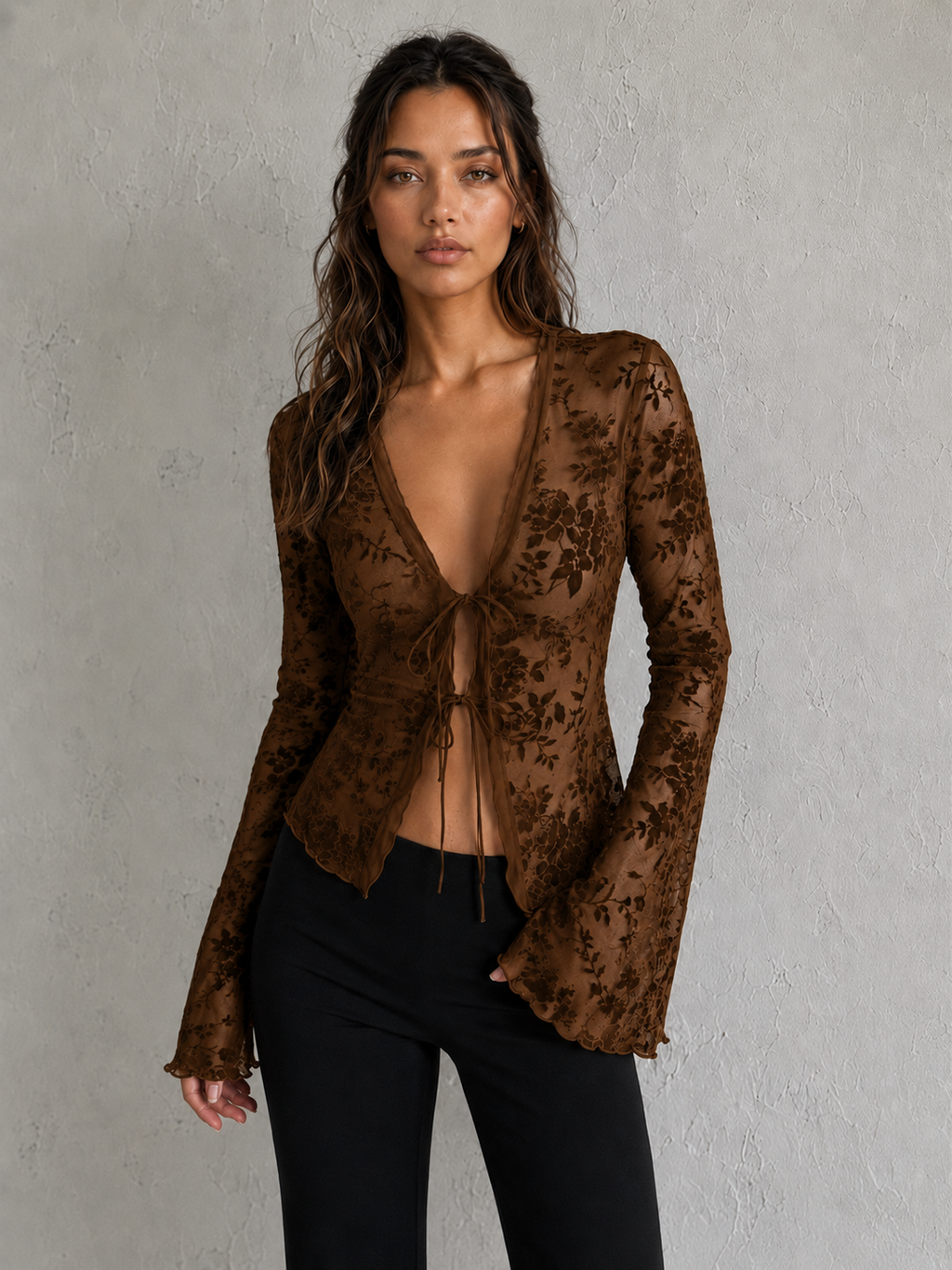 A woman modeling a brown floral lace long-sleeve wrap top with a deep V-neck and front tie closure, paired with black pants, against a textured gray wall.