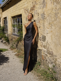 A woman wearing the Elévaison Amelie black one-shoulder satin maxi dress with a high slit, viewed from the front, beside a rustic stone wall.
