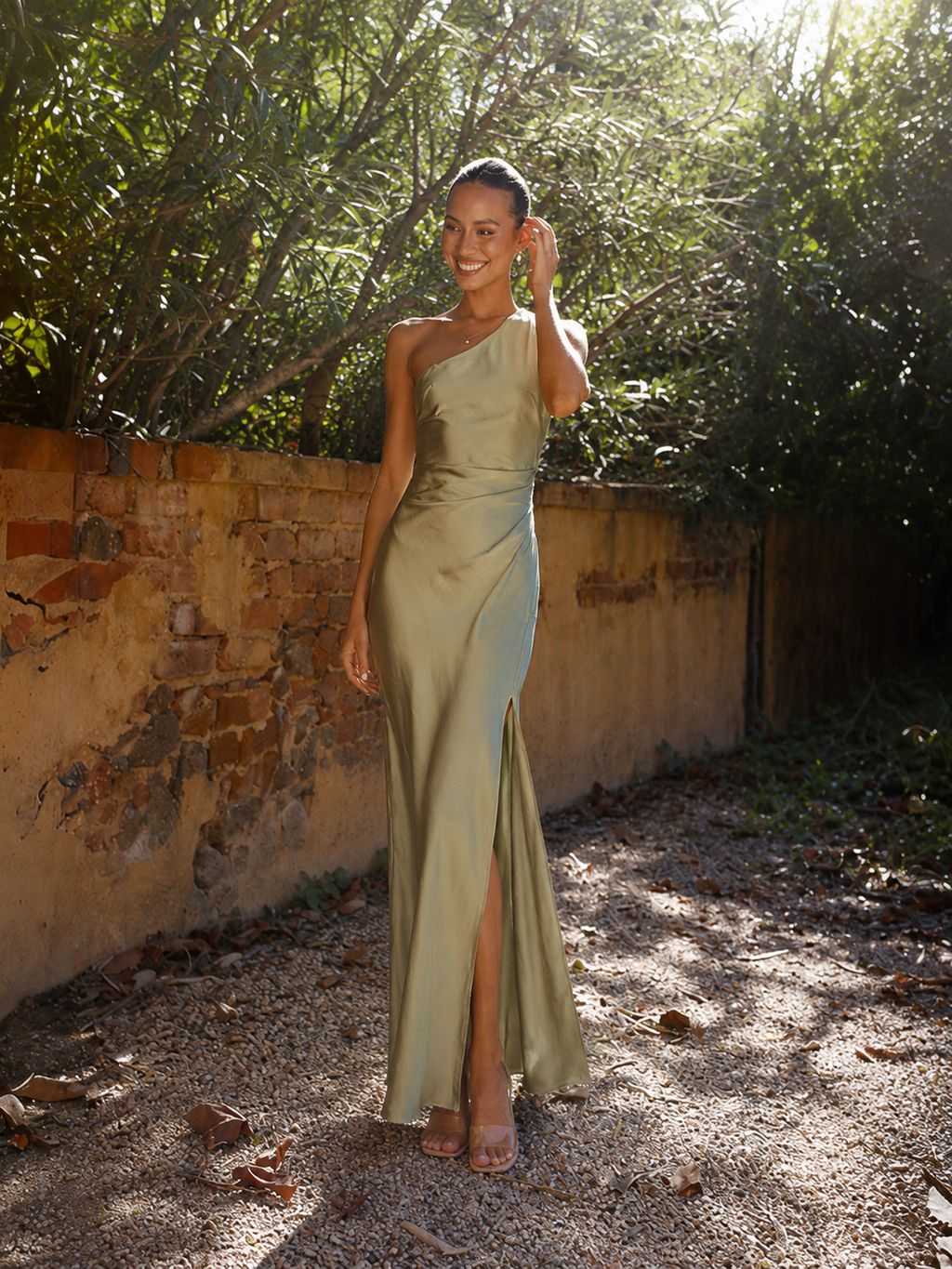 A woman wearing the Elévaison Amelie light green one-shoulder satin maxi dress with a high slit, viewed from the front, beside a garden wall.