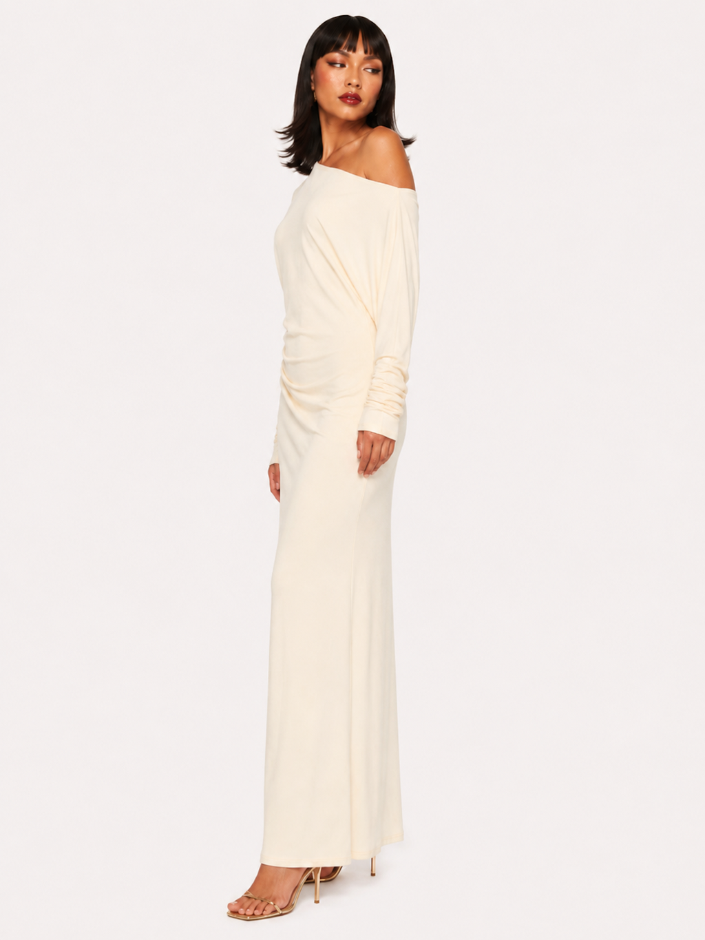 A woman viewed from the side wearing the Elévaison Arielle white asymmetric off-shoulder maxi dress, against an ivory studio background.
