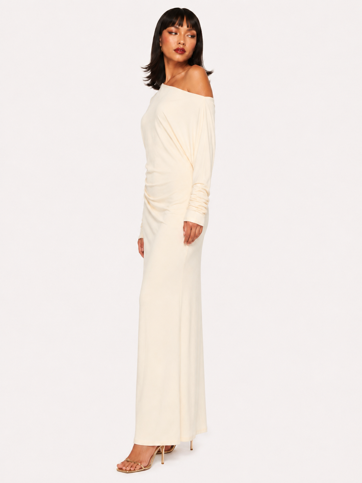 A woman viewed from the side wearing the Elévaison Arielle white asymmetric off-shoulder maxi dress, against an ivory studio background.