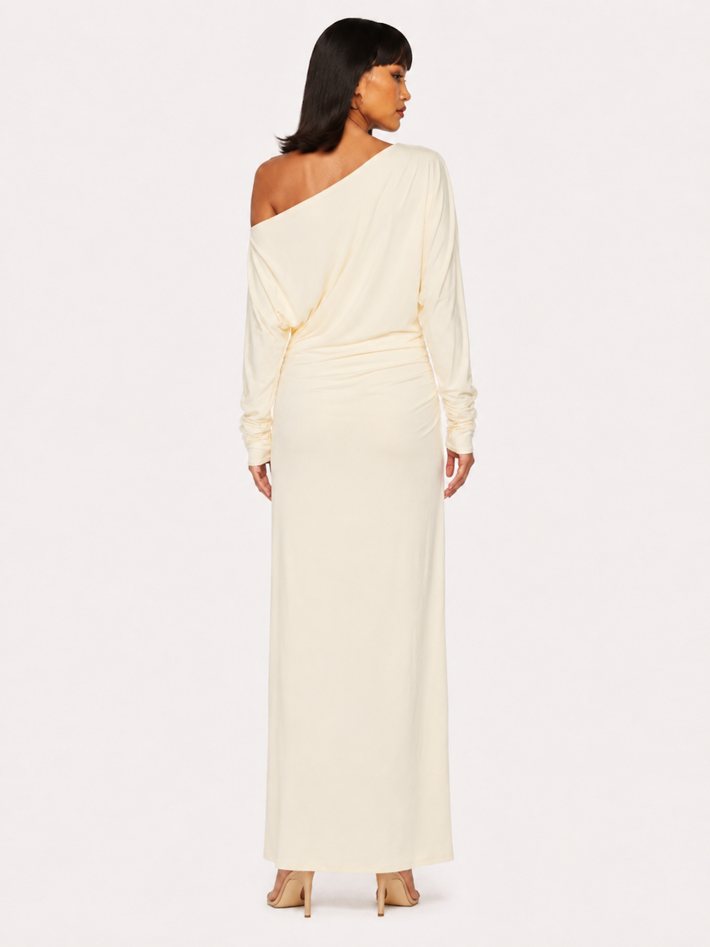A woman viewed from the back wearing the Elévaison Arielle white asymmetric off-shoulder maxi dress, against an ivory studio background.