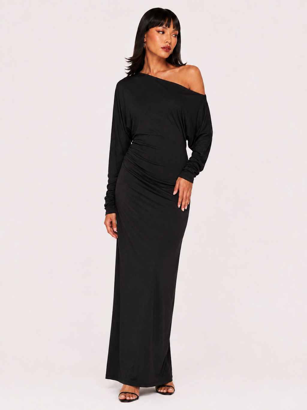 A woman wearing the Elévaison Arielle black asymmetric off-shoulder maxi dress, viewed from the front, against an ivory studio background.