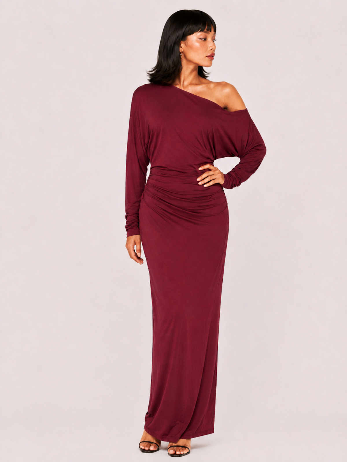 A woman wearing the Elévaison Arielle burgundy asymmetric off-shoulder maxi dress, viewed from the front, against a pale pink studio background.