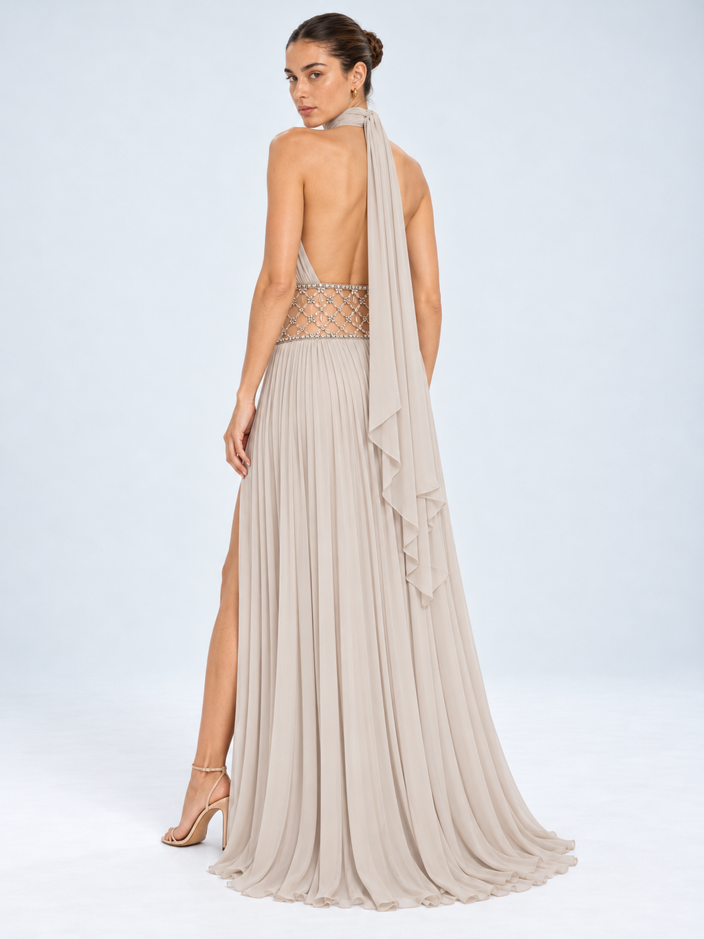A model shown from the back wearing a floor-length halter gown in a light taupe-beige chiffon. The dress features a sheer beaded waistband, a high leg slit, and a draped ruffled veil.