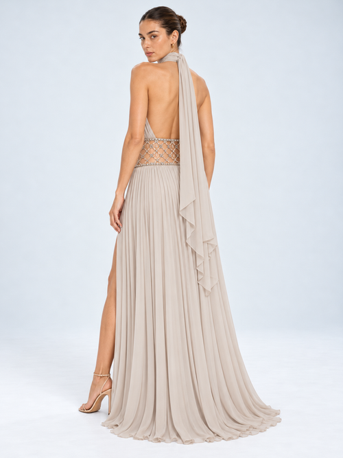 A model shown from the back wearing a floor-length halter gown in a light taupe-beige chiffon. The dress features a sheer beaded waistband, a high leg slit, and a draped ruffled veil.