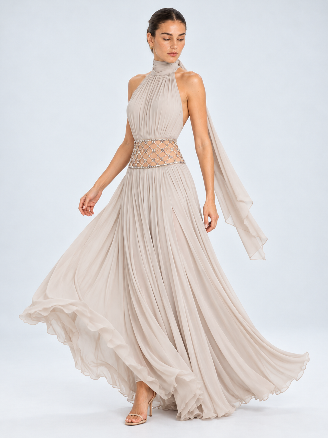 A woman modeling a floor-length halter gown in a light taupe-beige chiffon fabric with a beaded waistband and a flowing draped sleeve, photographed against a pale blue studio background.