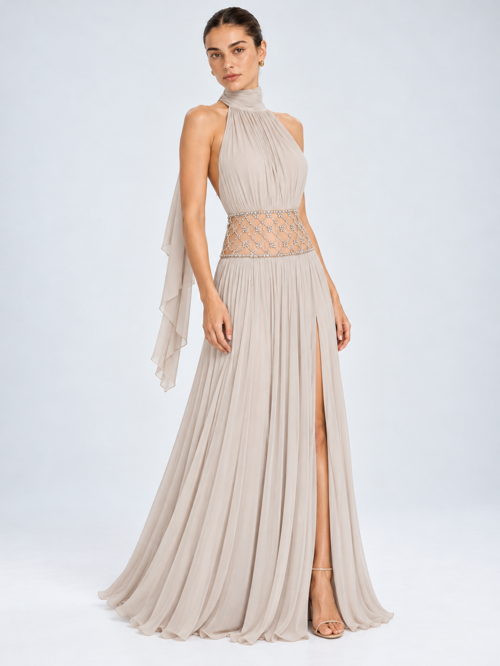 A woman models a floor-length halter gown in a light taupe-beige chiffon. The dress features a draped high neckline, a sheer beaded waistband, a flowing pleated skirt with a thigh-high slit, and a cascading sheer sleeve.