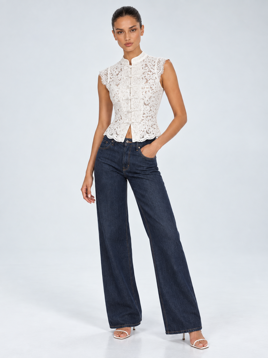Elévaison Isadora Blouse in white, front view showing a stand collar, frog-button front and scalloped lace hem, styled with dark wide-leg jeans against a pale blue studio background.