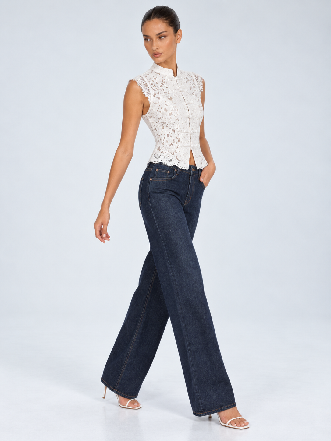 Elévaison Isadora Blouse in white, three-quarter view showing a stand collar, frog-button front and scalloped lace hem, styled with dark wide-leg jeans against a pale blue studio background.