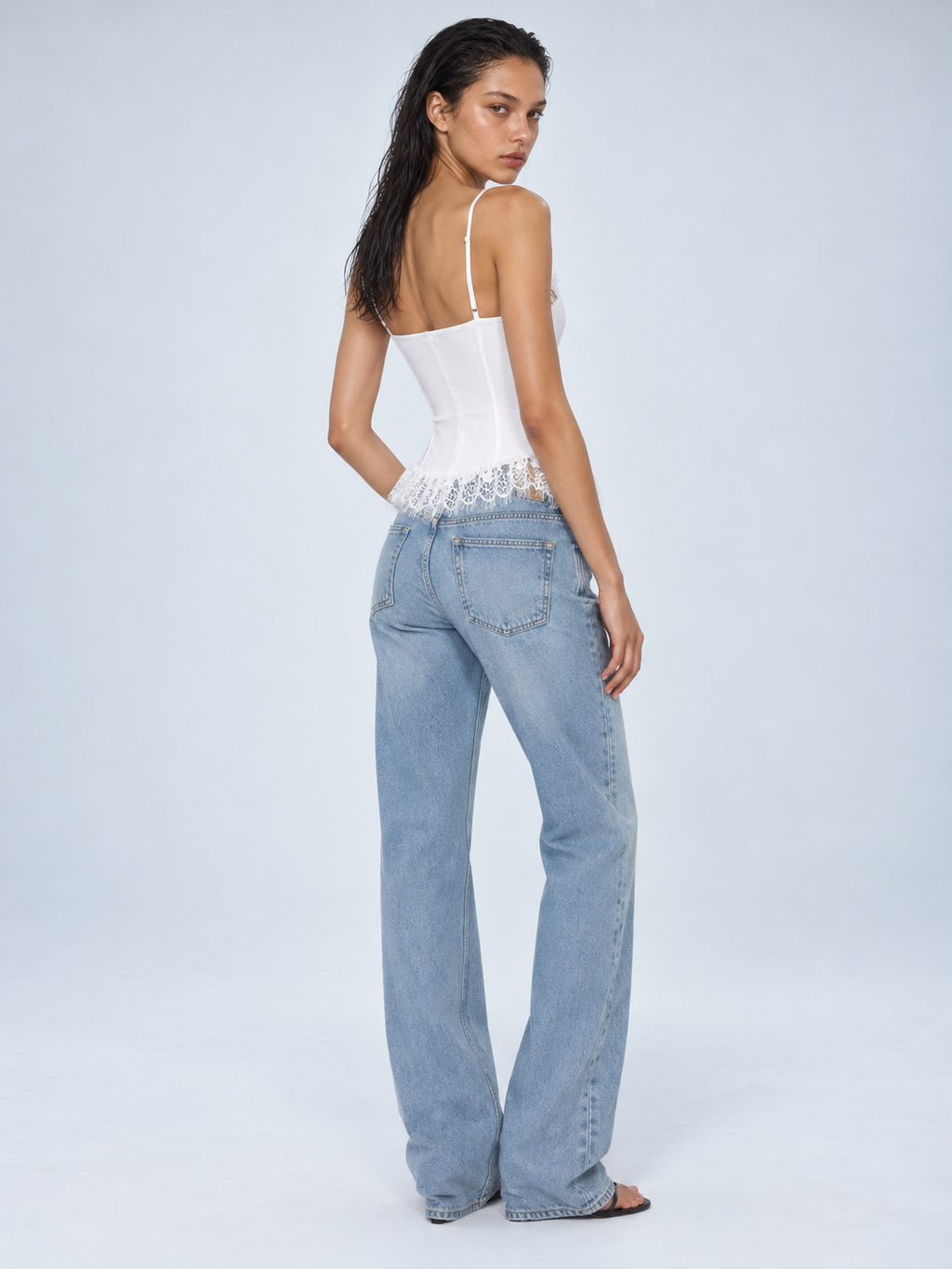 Elévaison Belle Tank Top in white, back view showing thin straps and a lace-trimmed peplum hem, styled with light blue jeans against a pale blue studio background.