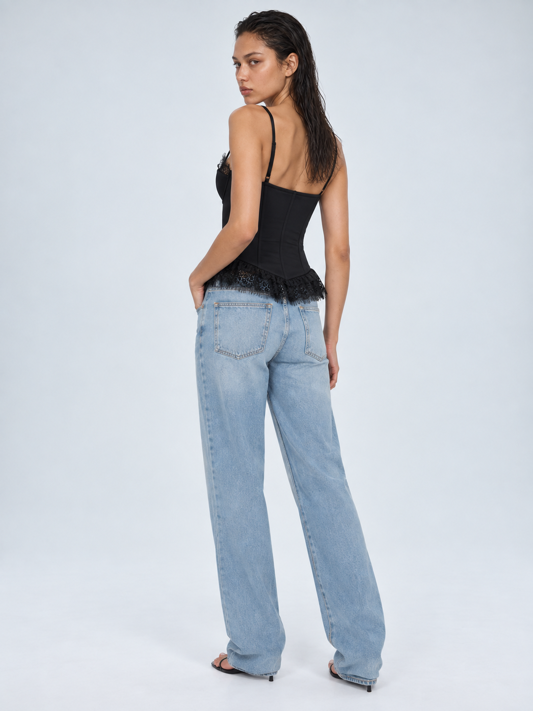 Elévaison Belle Tank Top in black, back view showing thin straps and a lace-trimmed peplum hem, styled with light blue jeans against a pale blue studio background.