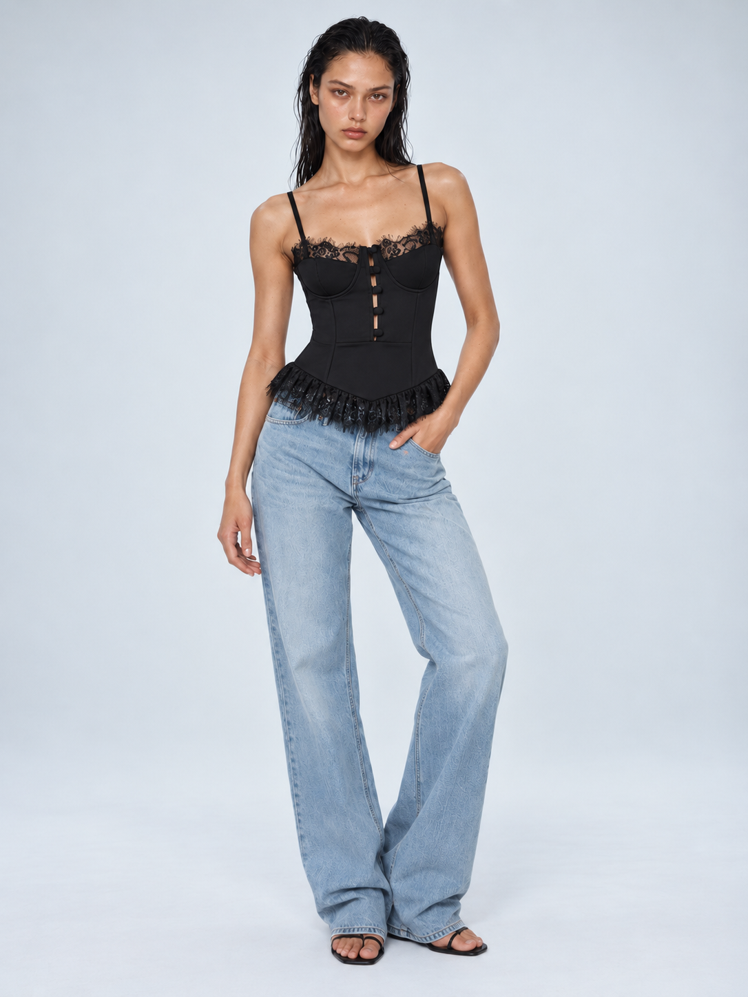 Elévaison Belle Tank Top in black, full-length front view showing thin straps, a fitted corset bodice and lace-trimmed hem, styled with light blue jeans against a pale blue studio background.