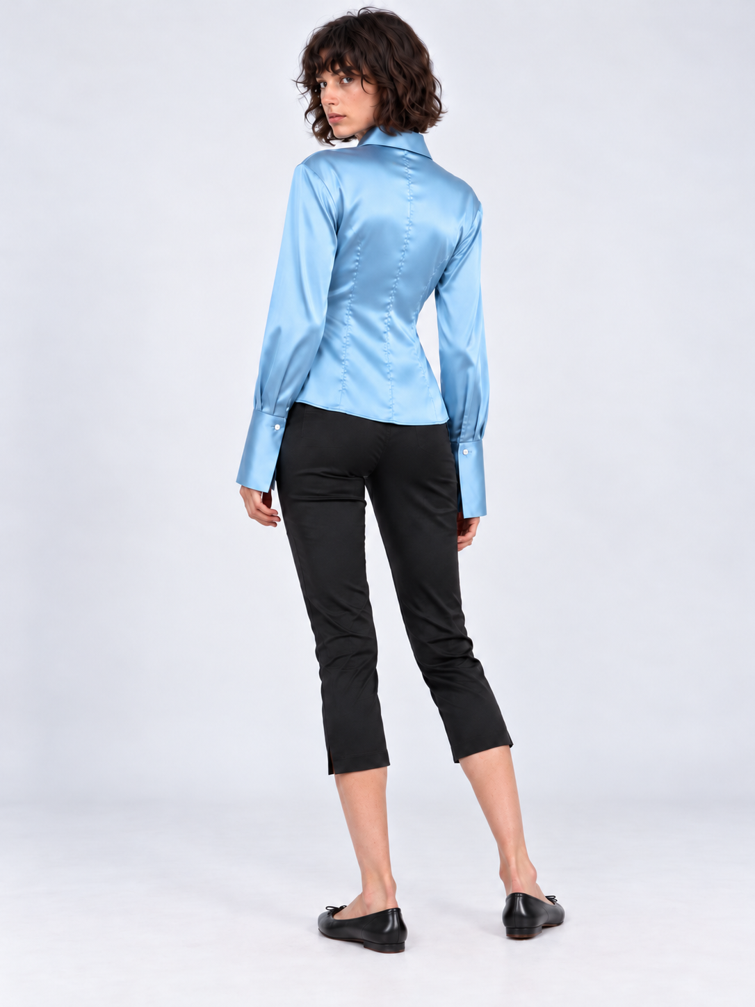 A model looks over her shoulder in the Elévaison Adrienne Shirt in Light Blue, showing its fitted back against a light gray backdrop.
