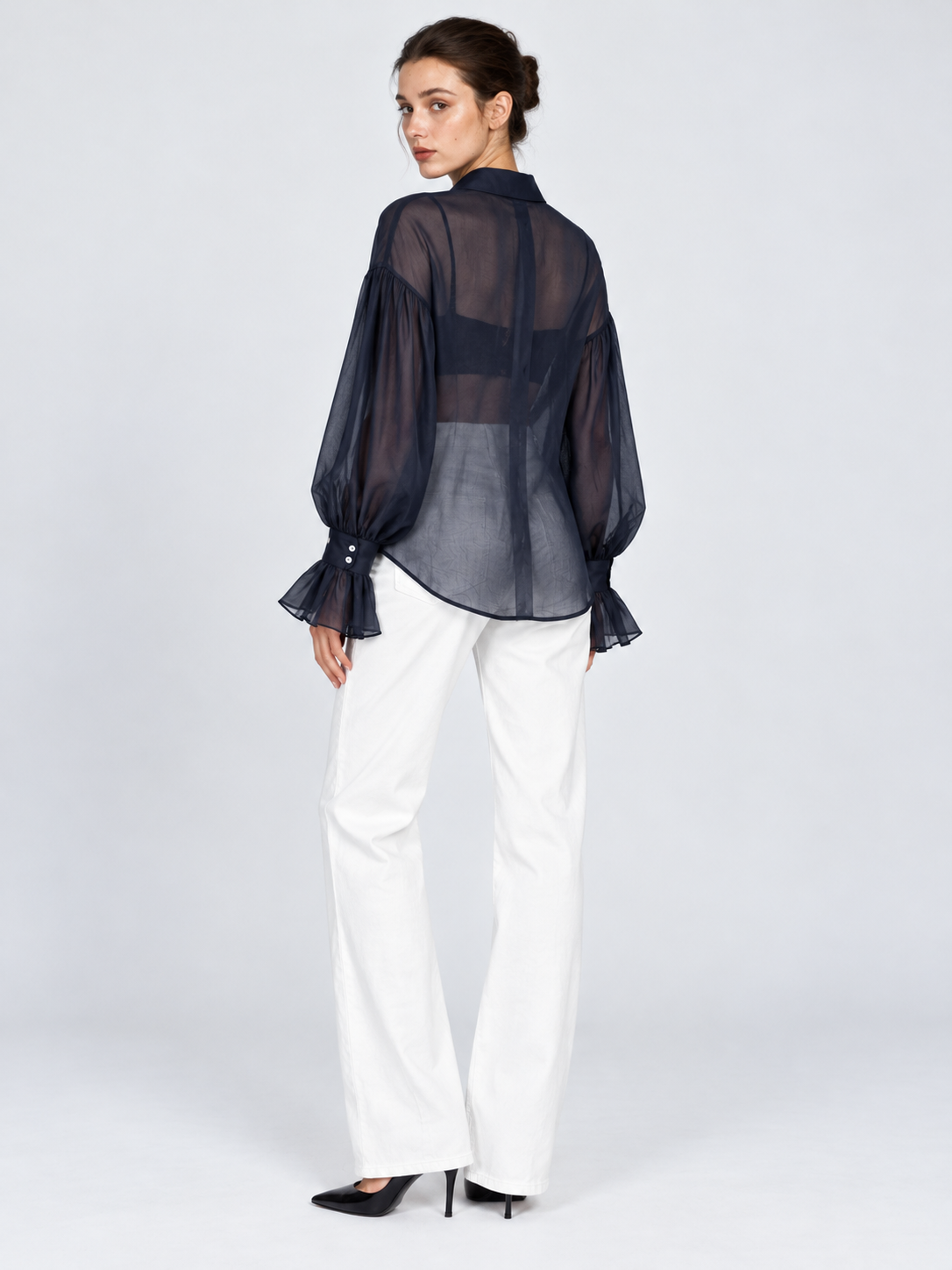 A model turns in the Elévaison Florence Blouse in Navy, revealing its sheer back and gathered sleeves against a light gray backdrop.