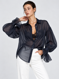 A model raises one arm in the Elévaison Florence Blouse in Navy, showing its full sleeve and ruffled cuff against a gray backdrop.
