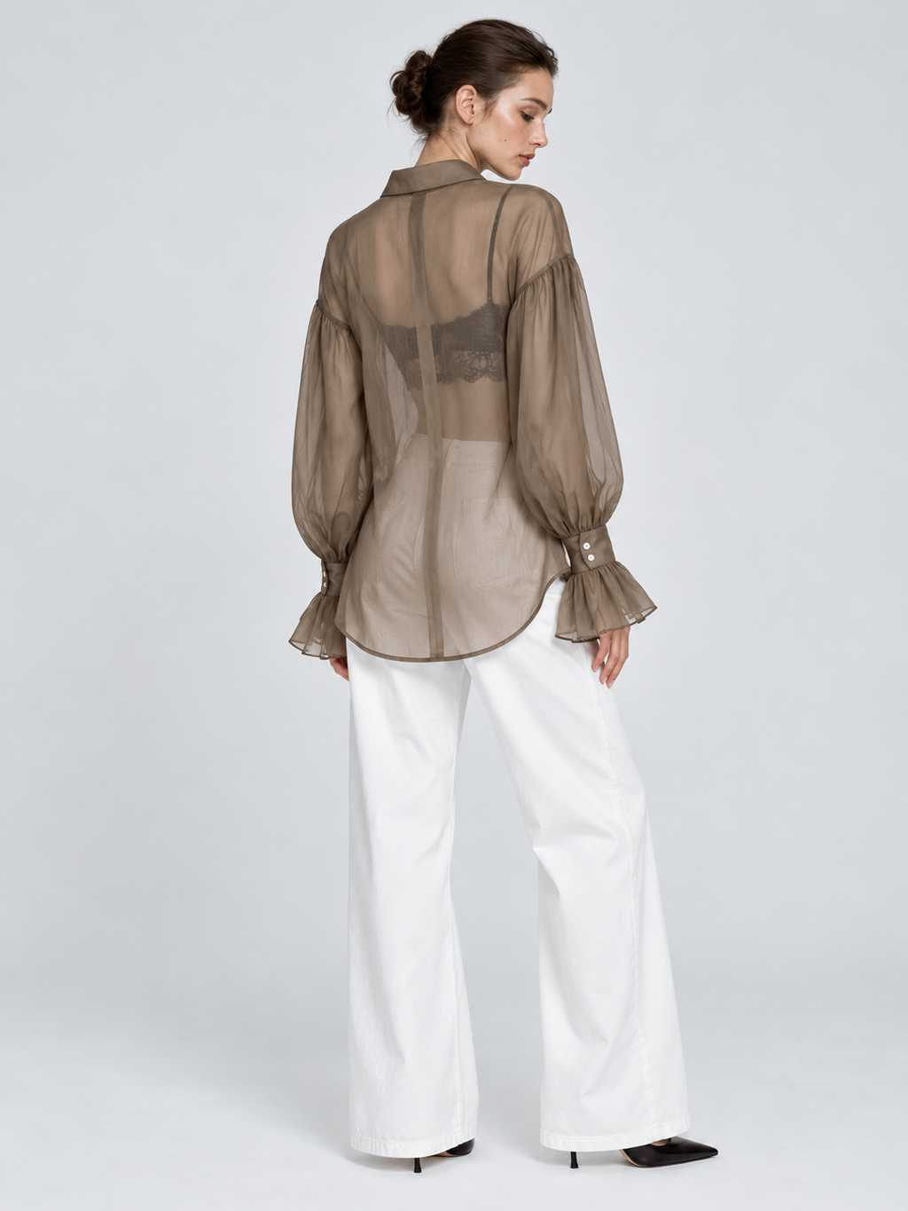A model looks back in the Elévaison Florence Blouse in Khaki, revealing its sheer back and curved hem against a light gray backdrop.