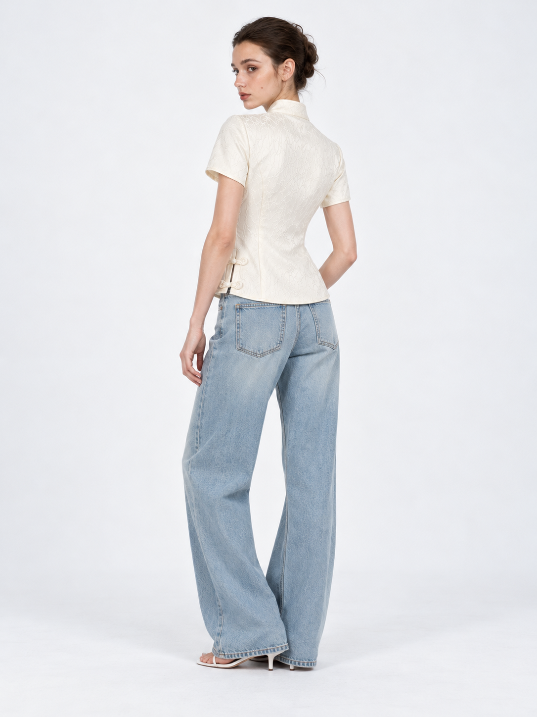 A model turns away in the Elévaison Beatrice Blouse in White, showing its fitted back and short sleeves against a light gray backdrop.
