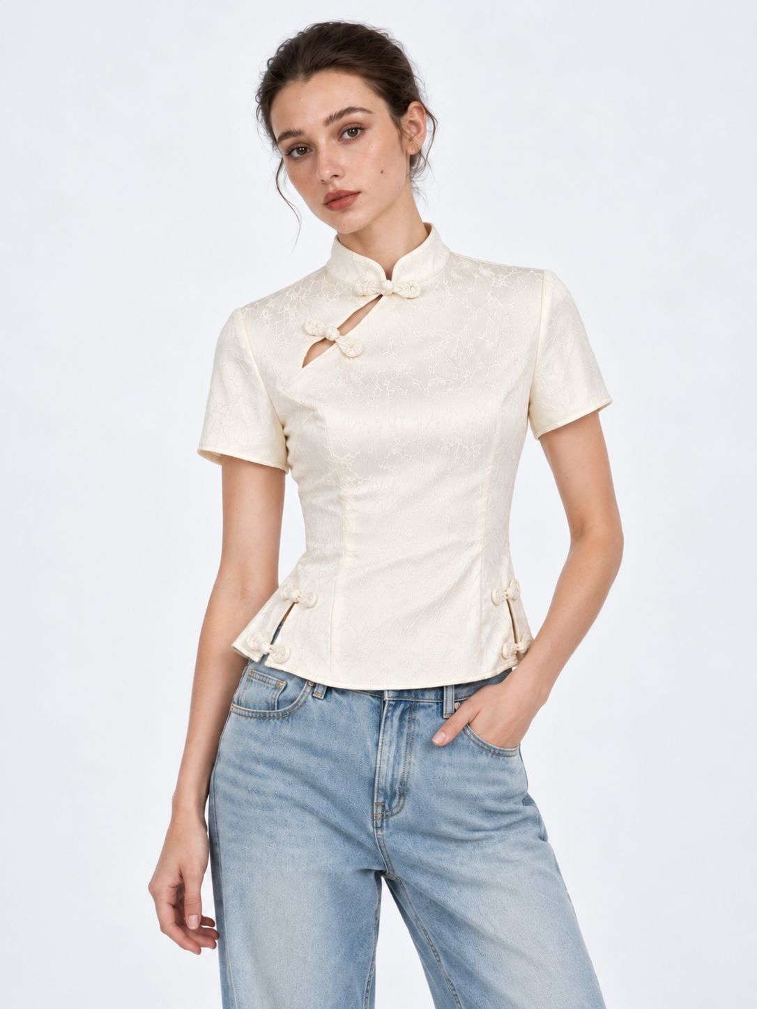 A model leans slightly in the Elévaison Beatrice Blouse in White, showing its subtle pattern and fitted shape against a gray backdrop.