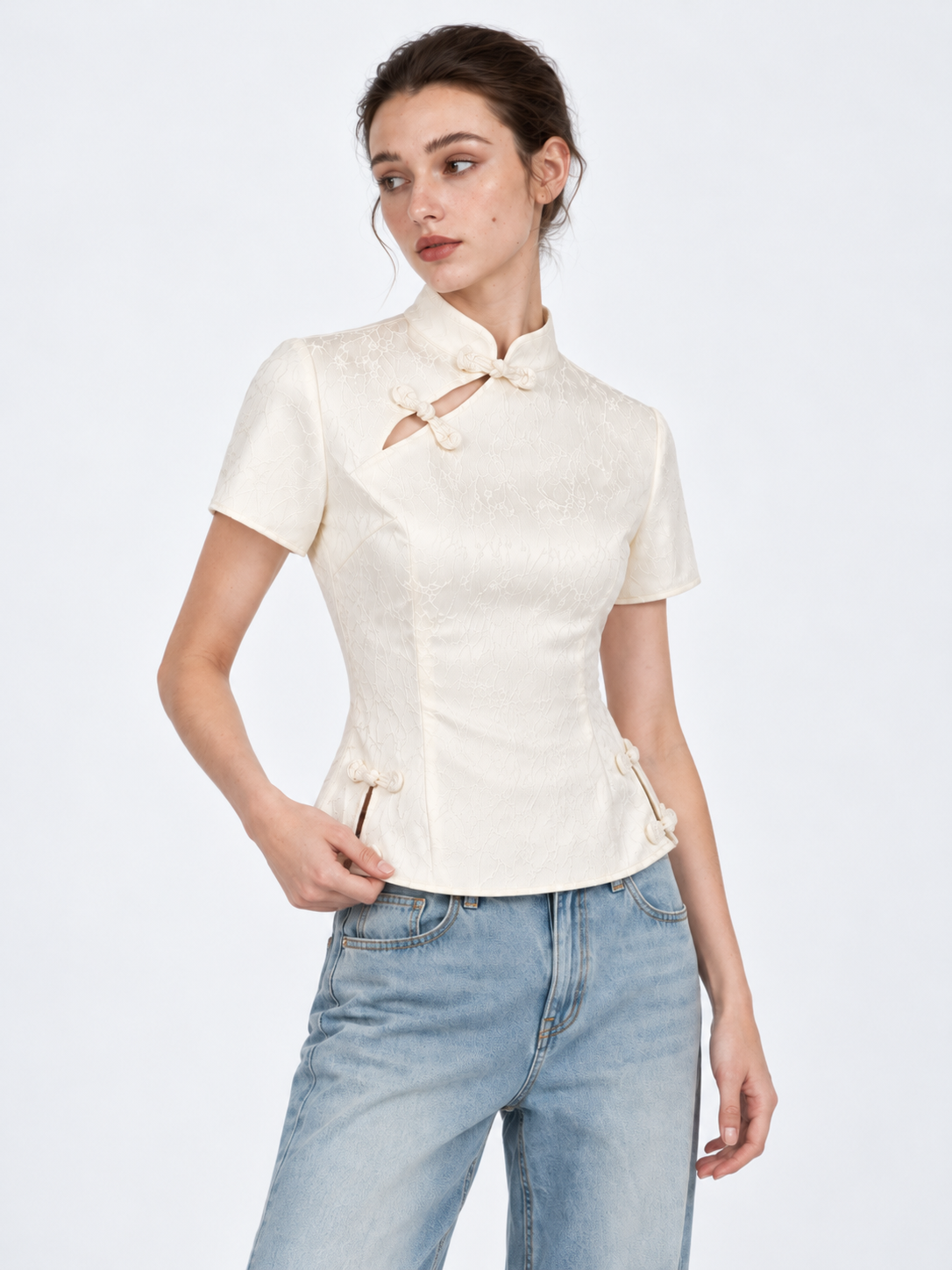 A model holds the hem of the Elévaison Beatrice Blouse in White, showing its side openings against a light gray studio backdrop.