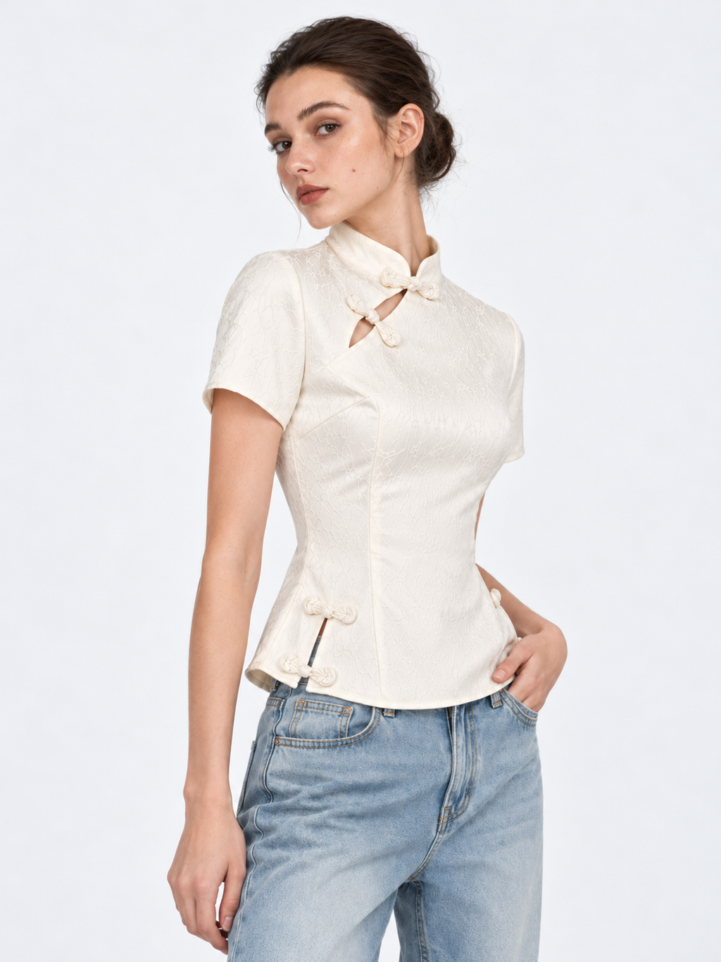An angled view of the Elévaison Beatrice Blouse in White shows its short sleeves and decorative closures against a gray backdrop.