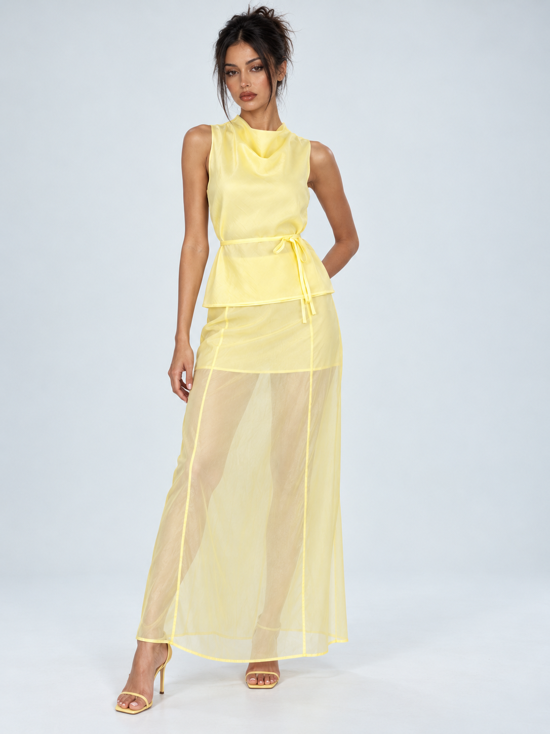 A model wears the Elévaison Alessia Two Piece in Yellow, with a sleeveless top and sheer long skirt against a light gray backdrop.