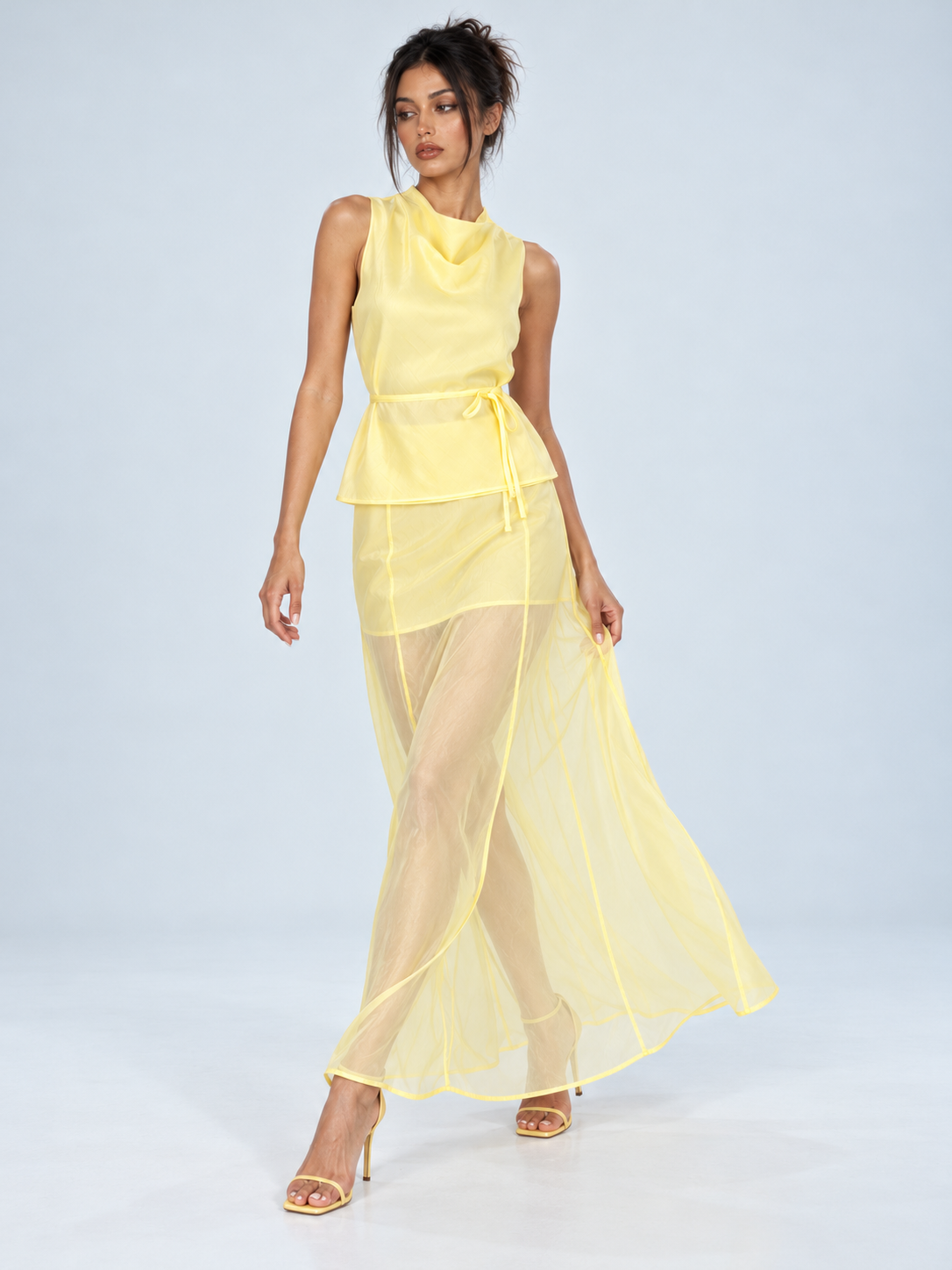 A model lifts the sheer skirt of the Elévaison Alessia Two Piece in Yellow, showing its flowing hem against a light gray backdrop.