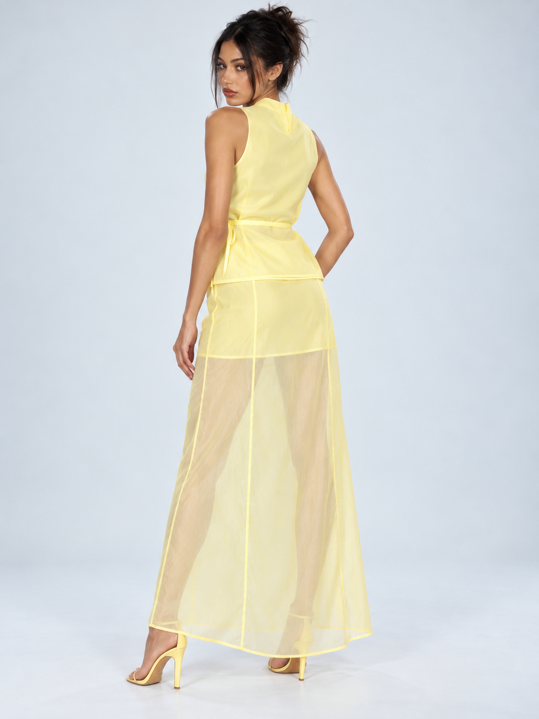 A model turns in the Elévaison Alessia Two Piece in Yellow, showing the back of the belted top against a light gray studio backdrop.