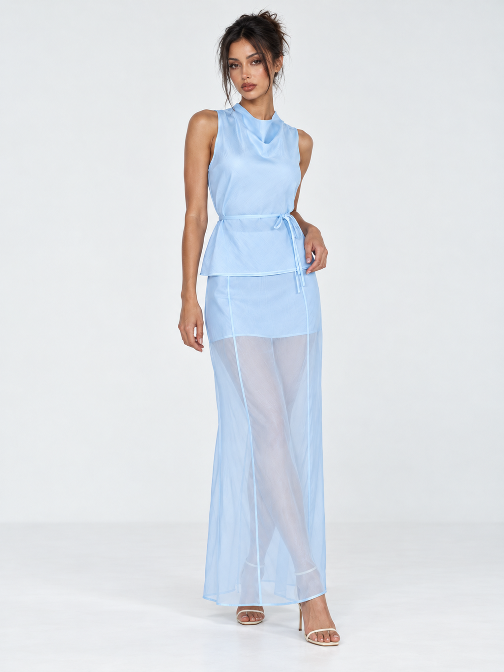 A model wears the Elévaison Alessia Two Piece in Blue, with a waist-tied top and sheer maxi skirt against a light gray backdrop.