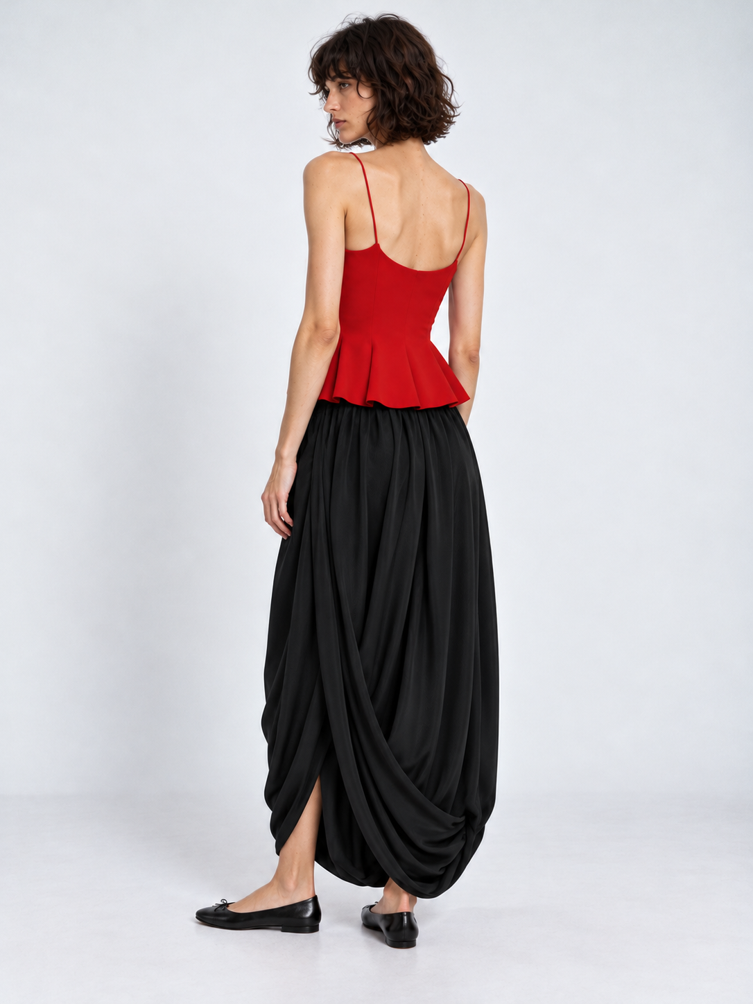 A model turns in the Elévaison Gabrielle Skirt in Black, showing the gathered back and draped hem against a light gray backdrop.