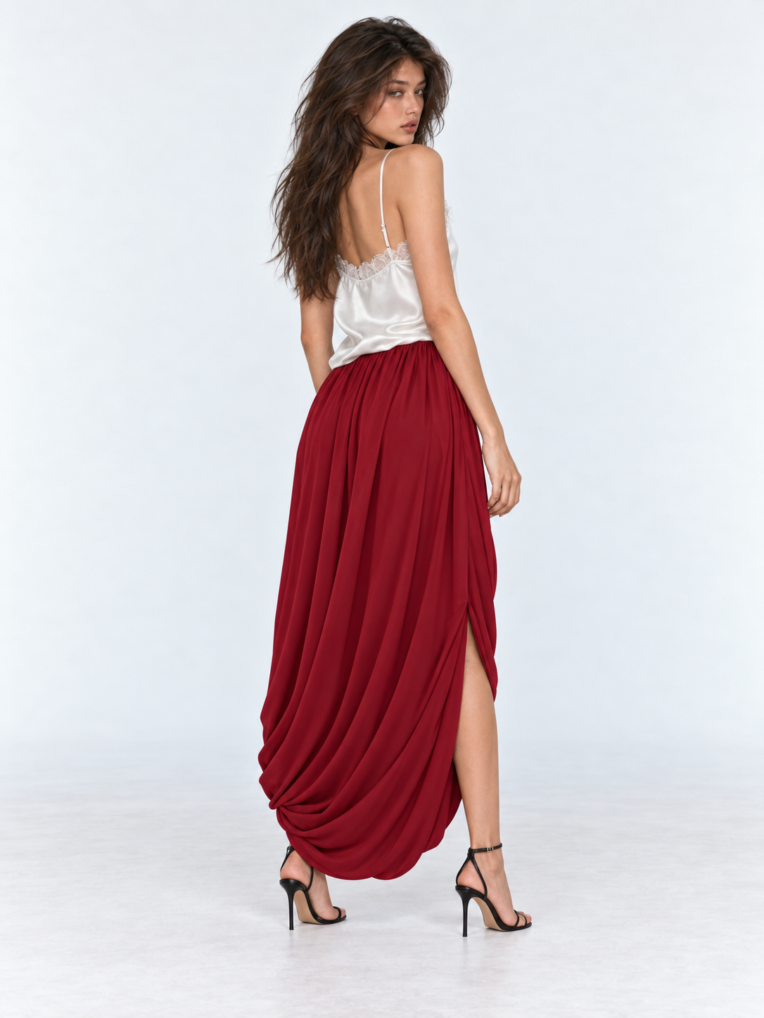 A model looks back in the Elévaison Gabrielle Skirt in Burgundy, showing its side opening and deep drapes against a gray backdrop.