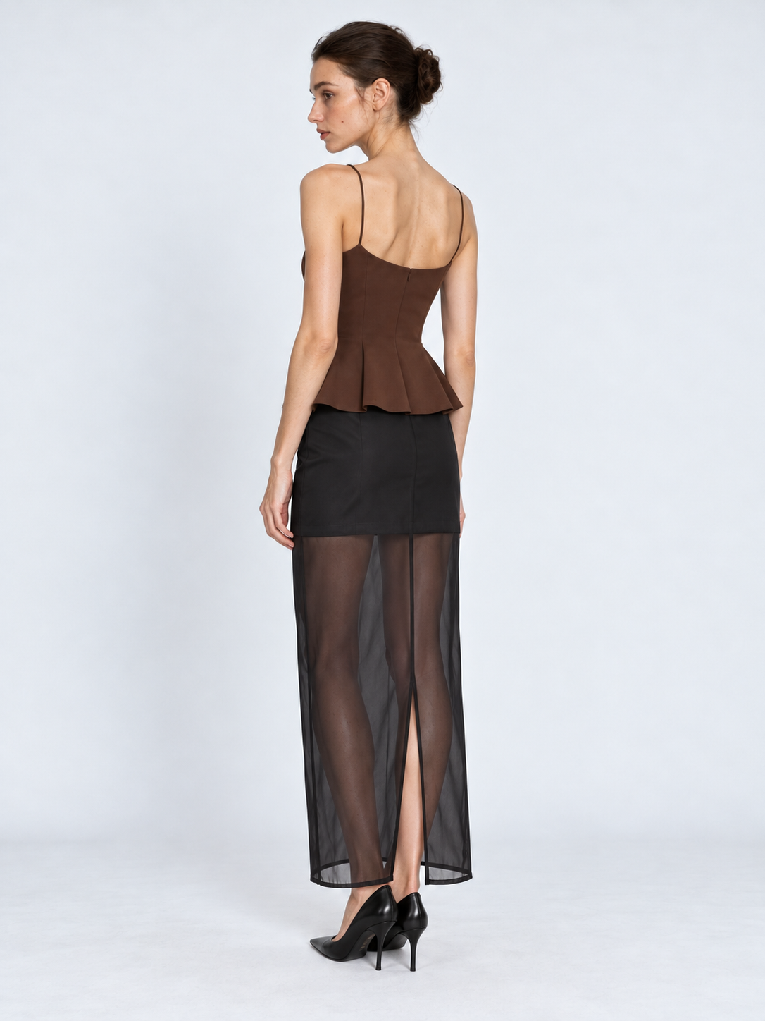 A model turns in the Elévaison Simone Skirt in Black, revealing the back slit and sheer maxi hem against a light gray backdrop.