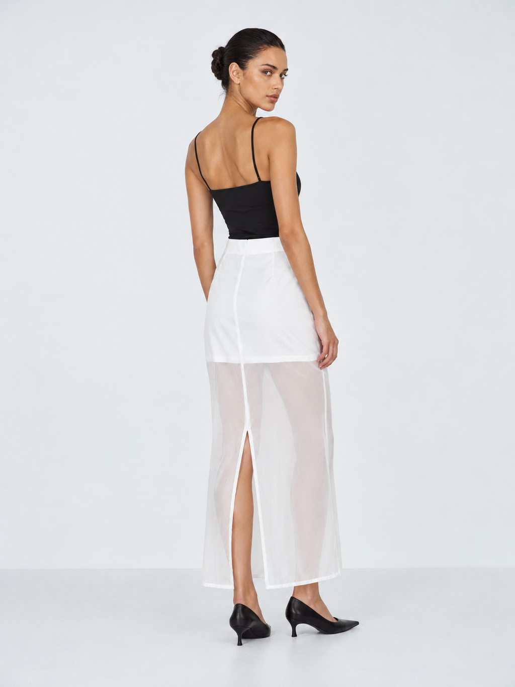 A model looks back in the Elévaison Simone Skirt in White, revealing the slit in its sheer long layer against a light gray backdrop.