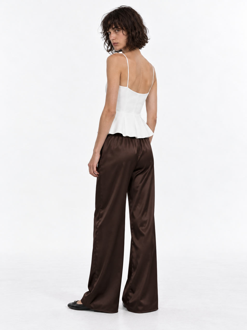 A model turns in the Elévaison Helena Pants in Brown, showing their wide legs and smooth back against a light gray studio backdrop.