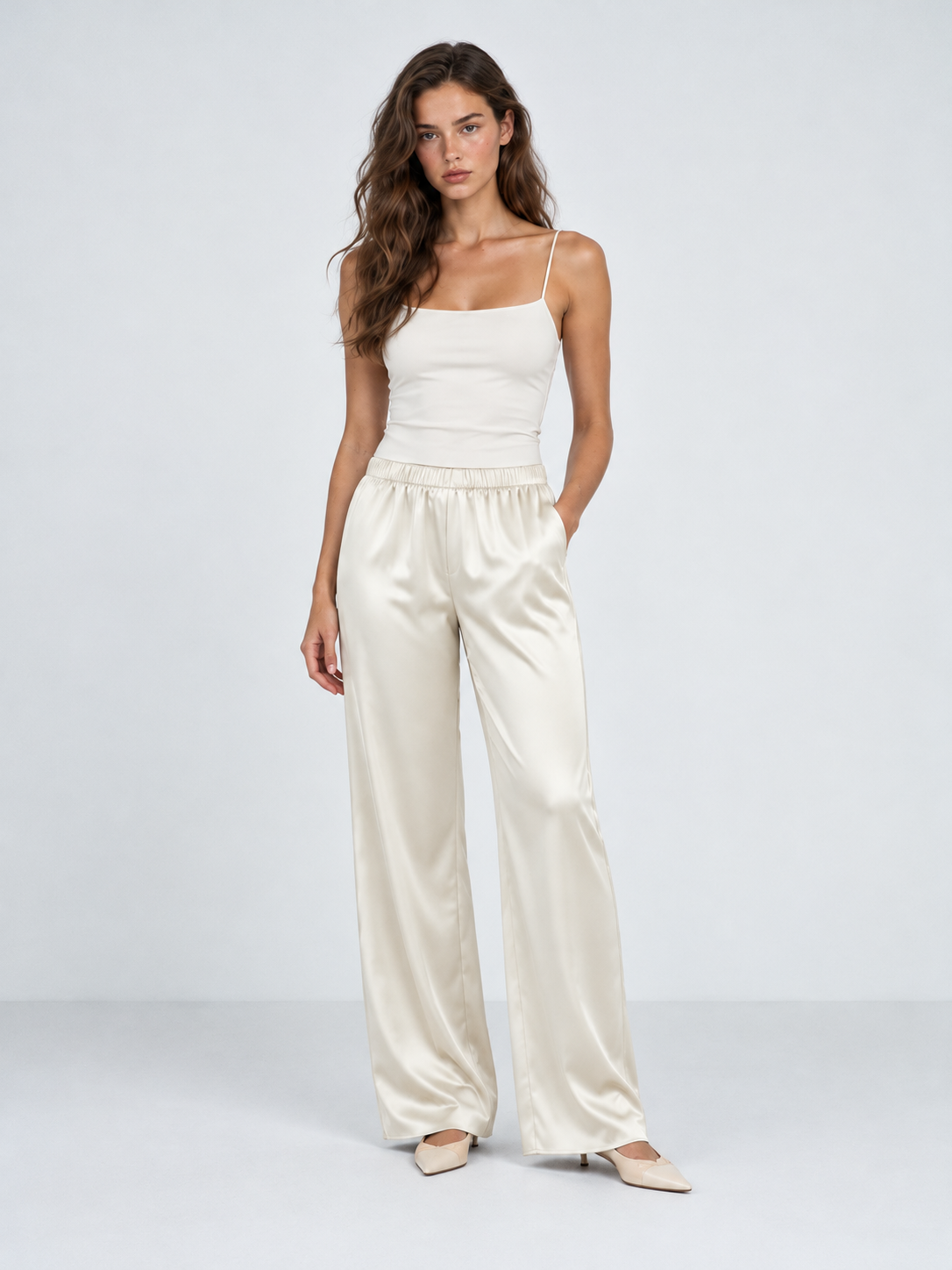 A model wears the Elévaison Helena Pants in Beige with one hand in a pocket, showing their wide legs against a light gray backdrop.