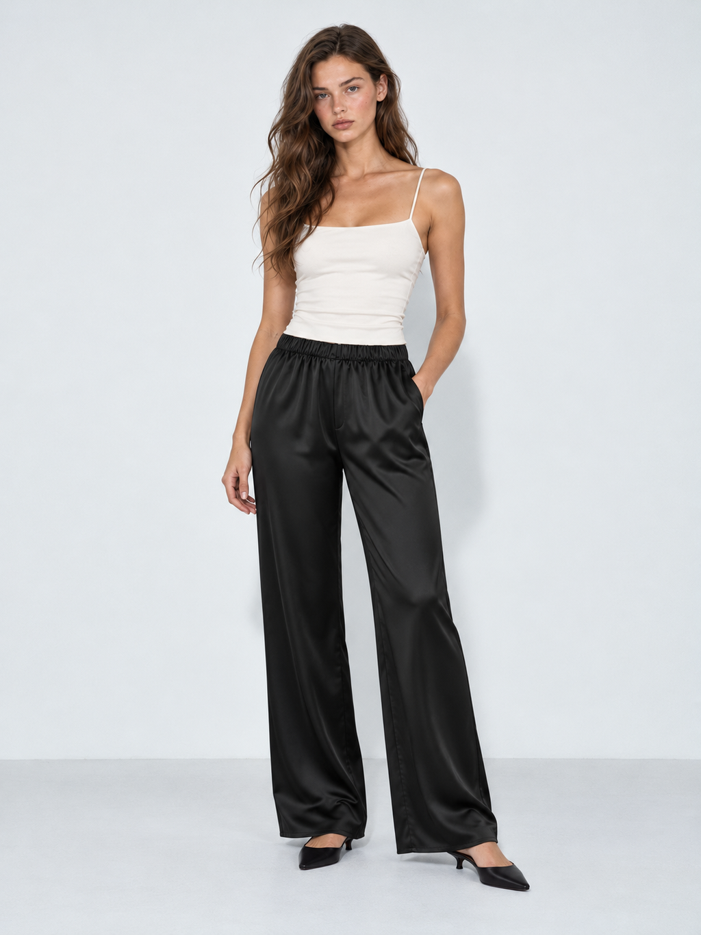 A model wears the Elévaison Helena Pants in Black, showing a gathered waist and satin finish against a light gray studio backdrop.