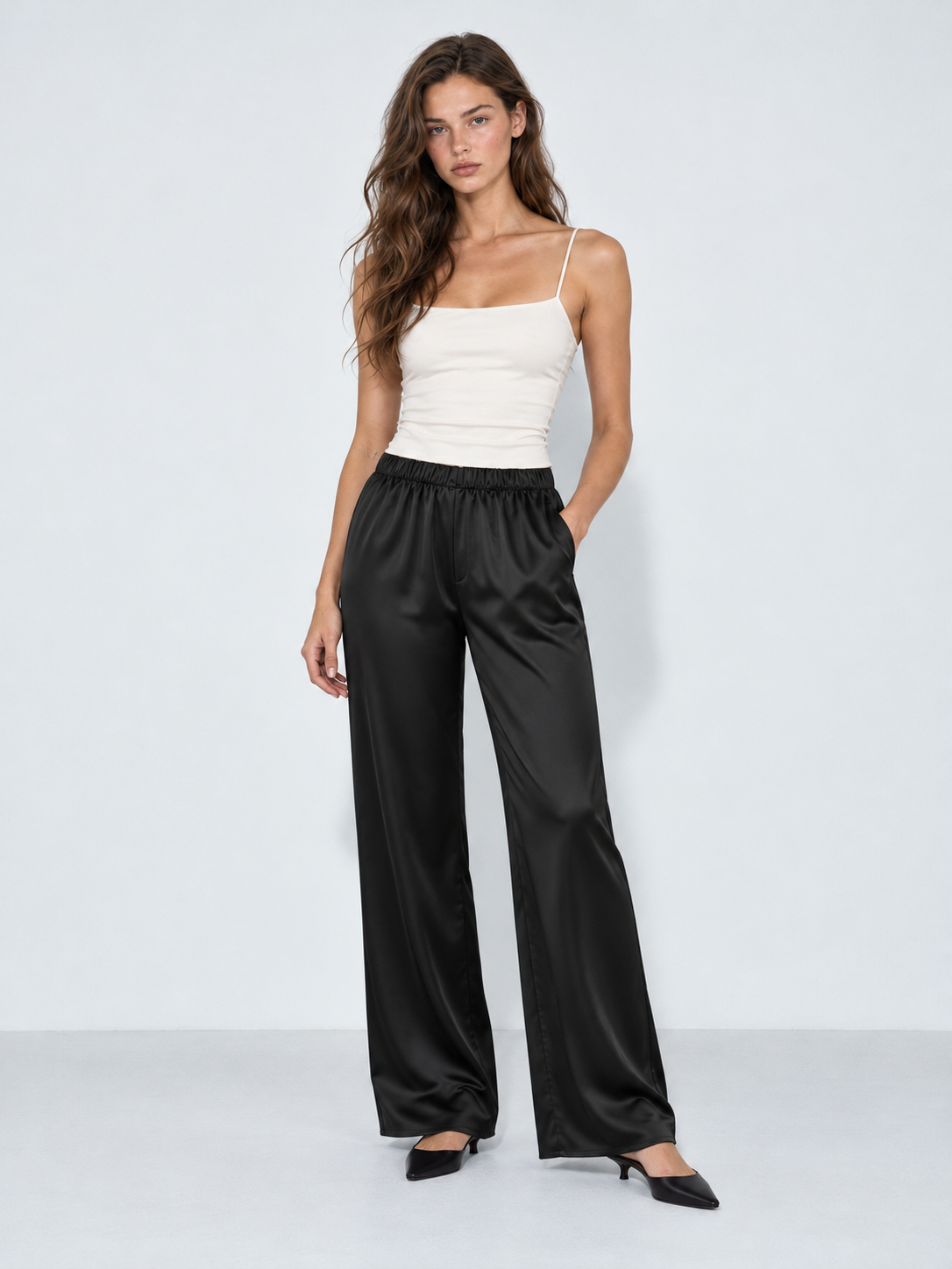 A model wears the Elévaison Helena Pants in Black, showing a gathered waist and satin finish against a light gray studio backdrop.