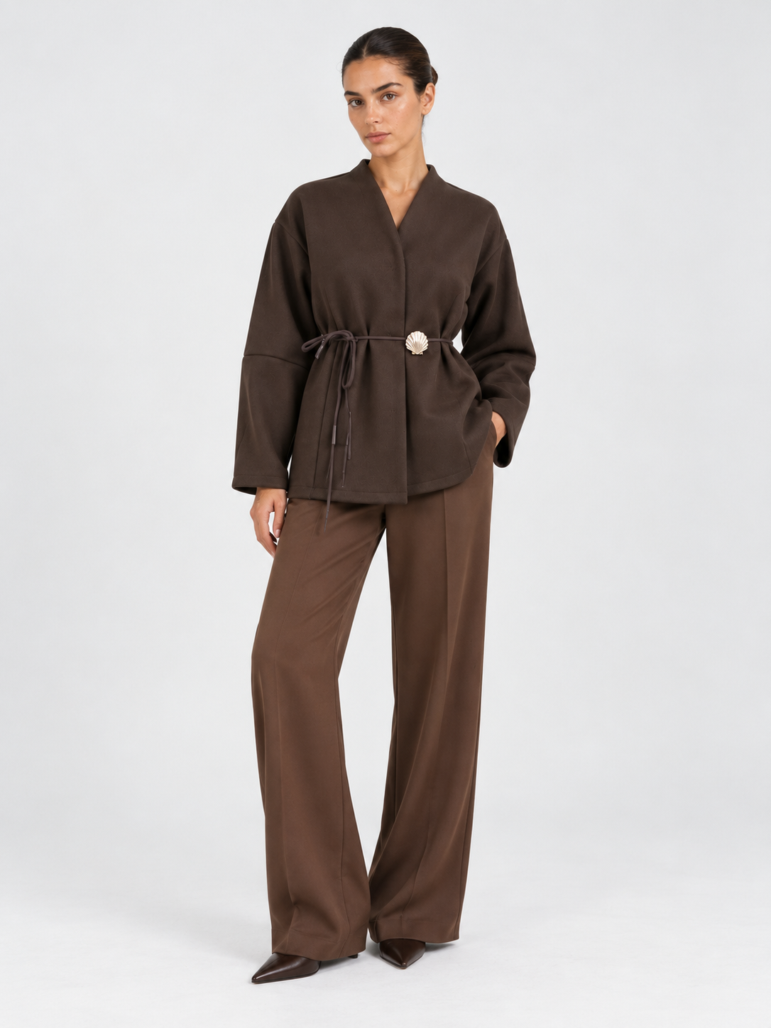 A model wears the Elévaison Freya Jacket in Brown, showing its tied waist and decorative clasp against a light gray studio backdrop.