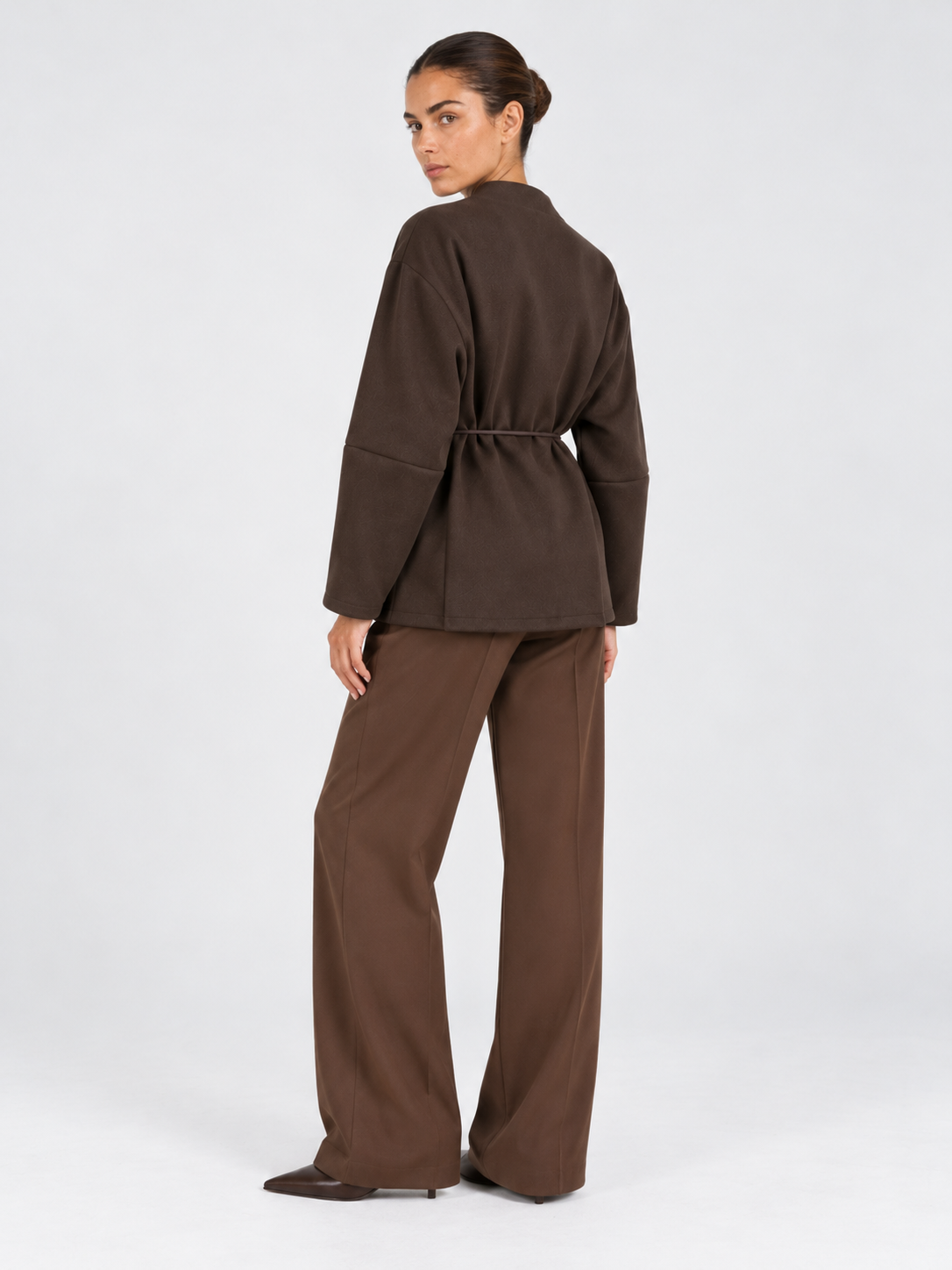 A model looks back in the Elévaison Freya Jacket in Brown, revealing its gathered waist and loose sleeves against a gray backdrop.