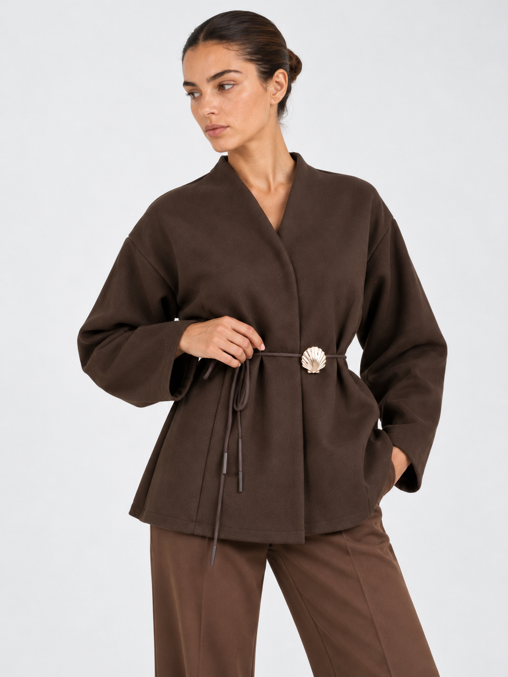 A closer front view of the Elévaison Freya Jacket in Brown shows its waist tie and flower-shaped clasp against a light gray backdrop.