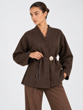 A closer front view of the Elévaison Freya Jacket in Brown shows its waist tie and flower-shaped clasp against a light gray backdrop.