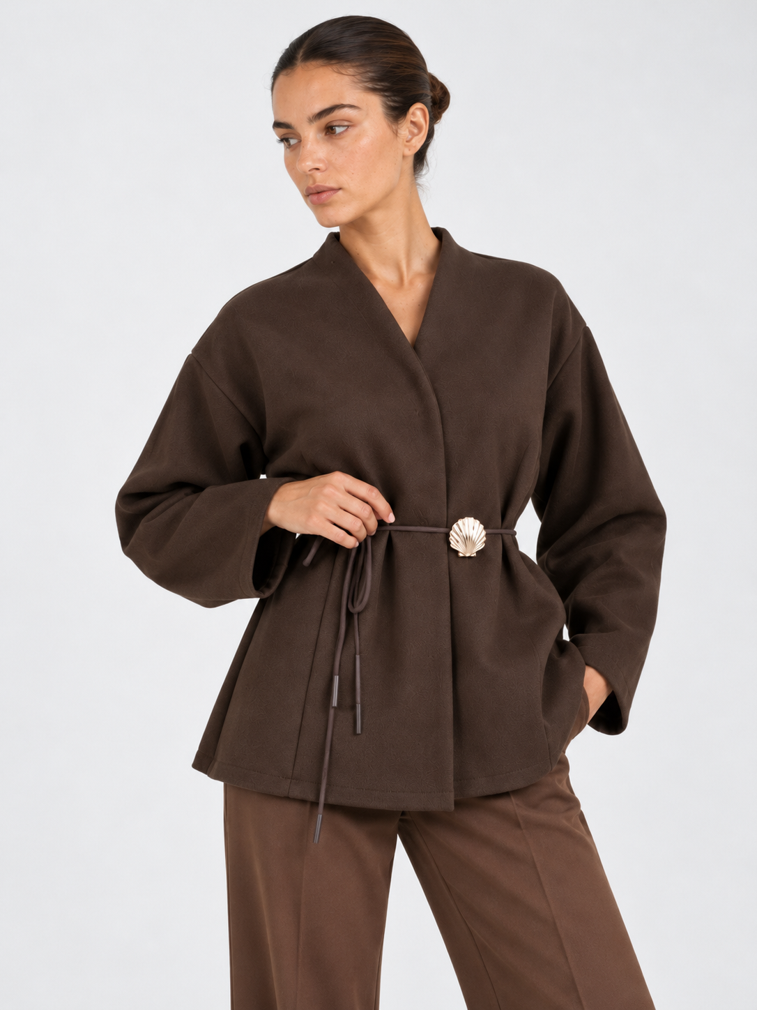 A closer front view of the Elévaison Freya Jacket in Brown shows its waist tie and flower-shaped clasp against a light gray backdrop.