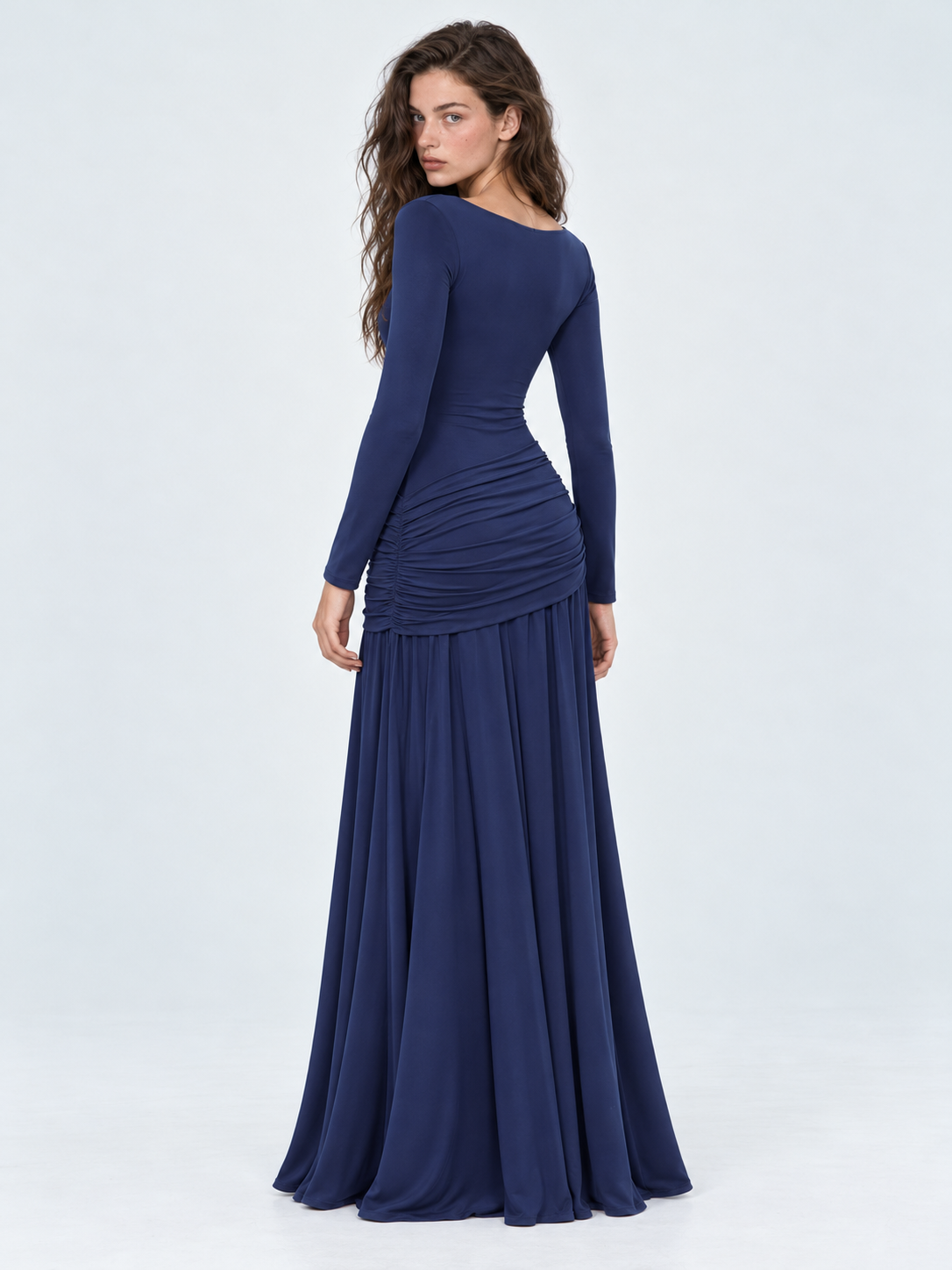 A model turns away in the Elévaison Arabella Dress in Dark Blue, showing its fitted back and gathered hips against a gray backdrop.
