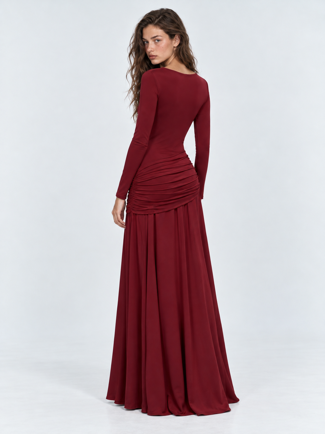 A model looks back in the Elévaison Arabella Dress in Burgundy, showing its fitted back and long skirt against a light gray backdrop.