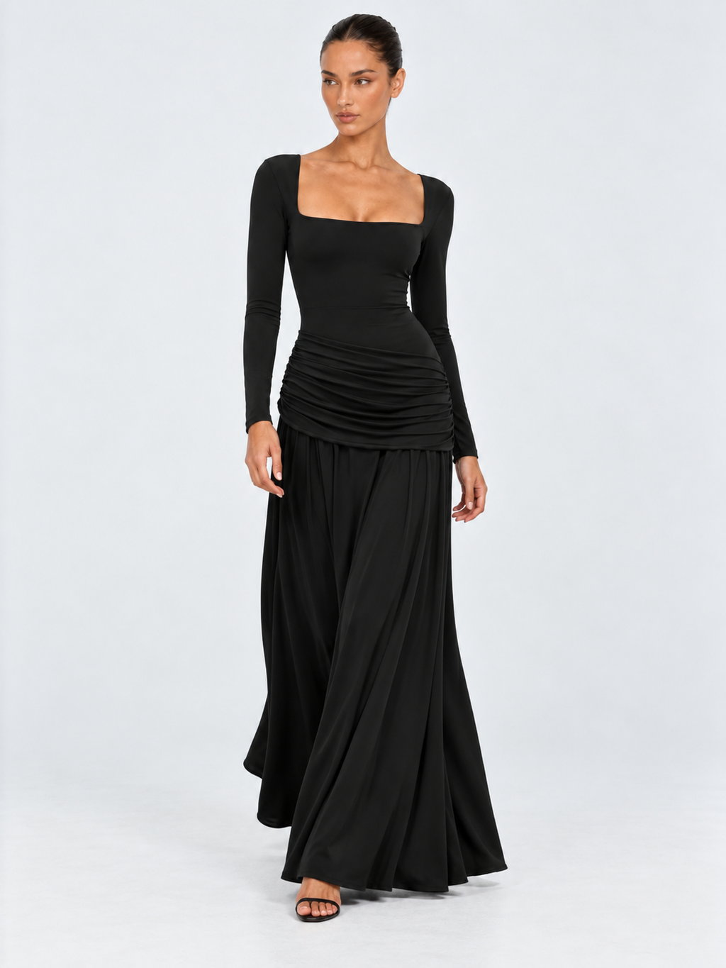 A model steps forward in the Elévaison Arabella Dress in Black, showing its flowing hem and ruched hips against a light gray backdrop.