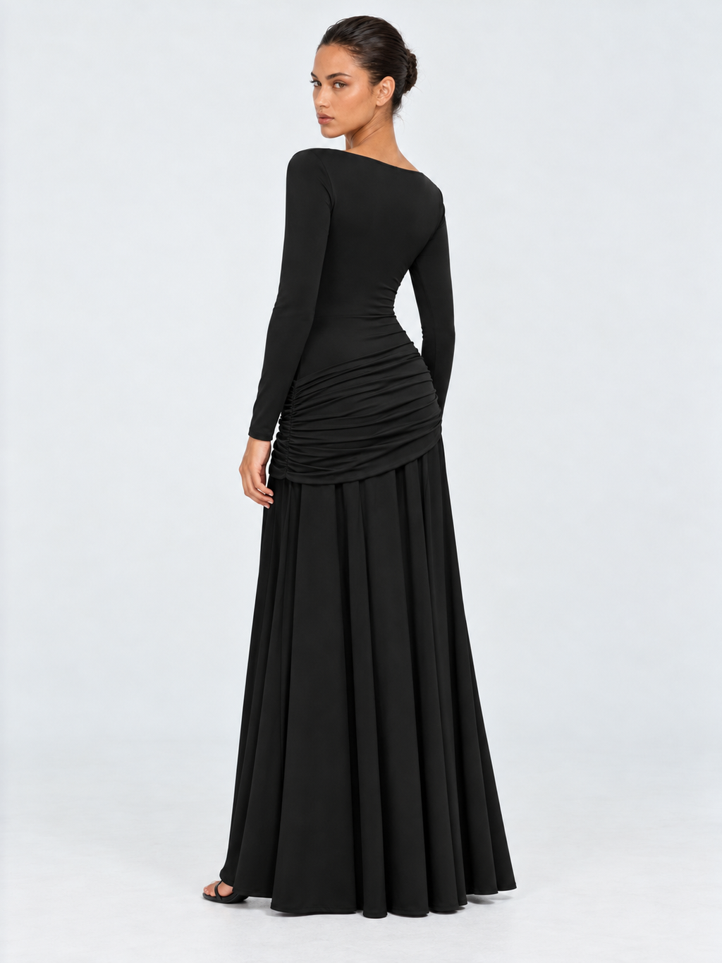 A model turns in the Elévaison Arabella Dress in Black, showing its fitted back and gathered hip panel against a light gray backdrop.