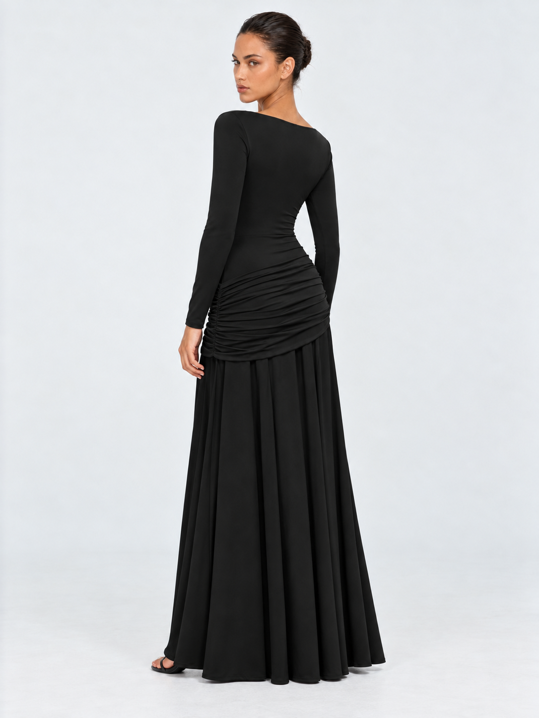 A model turns in the Elévaison Arabella Dress in Black, showing its fitted back and gathered hip panel against a light gray backdrop.